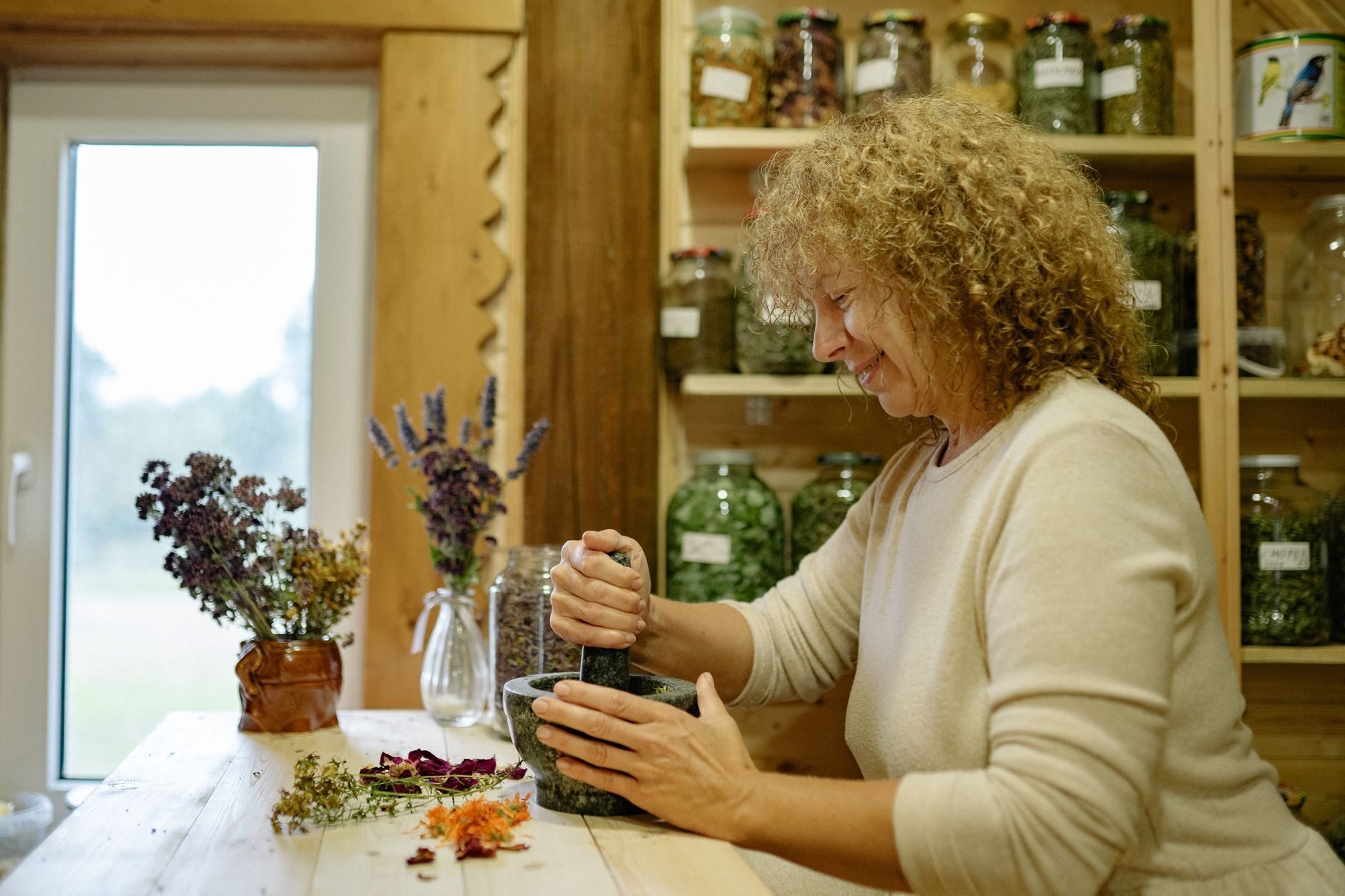 Herbalist grinding herbs before a wall of labeled apothecary jars